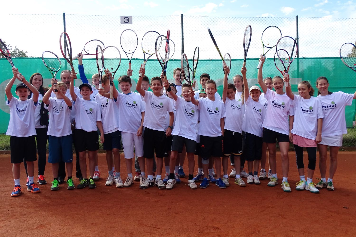 25 Mädchen und Jungen in weißen Tennis-Shirts halten lachend ihre Schläger in die Luft - Sommertraining im TSV Starnberg